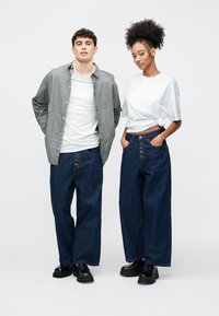 Δύο μοντέλα φορούν καθημερινά ρούχα: μία ριγέ πουκαμίσα με κουμπιά, λευκό μπλουζάκι και άνετα σκούρα μπλε τζιν. Το τζιν έχει κούμπωμα με κουμπιά.