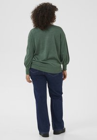 Grön stickad tröja med avslappnad passform och långa ärmar, i kombination med mörkblå denimjeans. Texturen är mjuk med en subtil glans.