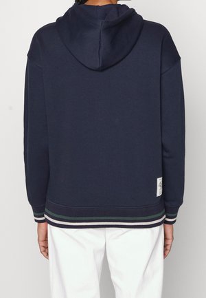 Personne portant un sweat à capuche bleu marine avec des poignets et un ourlet rayés, debout de dos, associée à un pantalon blanc.