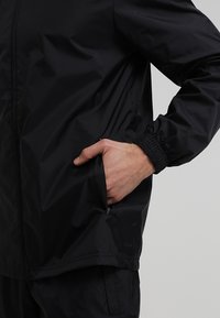 Henkilö, jolla on päällään musta pitkähihainen takki, käsi sivutaskussa, käsivarren joustava kuvio näkyvissä, vaalea harmaa tausta.