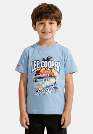 Jeune garçon souriant, portant un t-shirt bleu clair avec un motif de plage "Lee Cooper" et un pantalon noir, debout devant un fond uni.