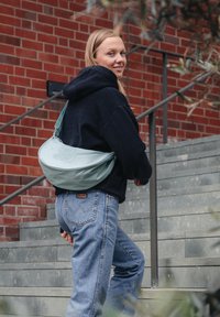 En mjuk, mintgrön axelväska med rund form och justerbar rem, som bärs över en marinblå huvtröja och blekta blå jeans.