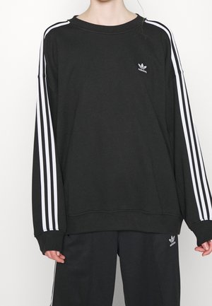 Personne portant un survêtement Adidas noir avec trois bandes blanches sur les manches et les jambes du pantalon, logo Adidas sur la poitrine et la cuisse.