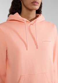 Felpa rosa chiaro con cappuccio regolabile, tessuto testurizzato e una piccola etichetta di marca sul petto. Presenta una tasca a marsupio sul davanti.
