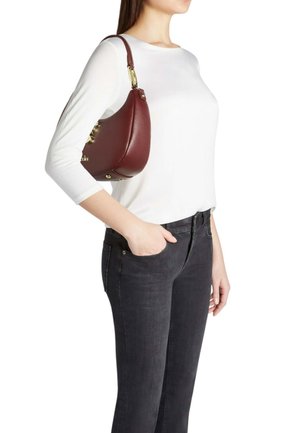 Borsa a mano in pelle color bordeaux con forma curva, dettagli in oro e manico corto, tenuta da una persona con maglia bianca e jeans scuri.