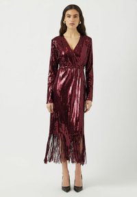 Abito lungo in paillettes bordeaux con scollatura a V, maniche lunghe, vita cin cinata e orlo frastagliato. Abbinato a scarpe nere con tacco alto.