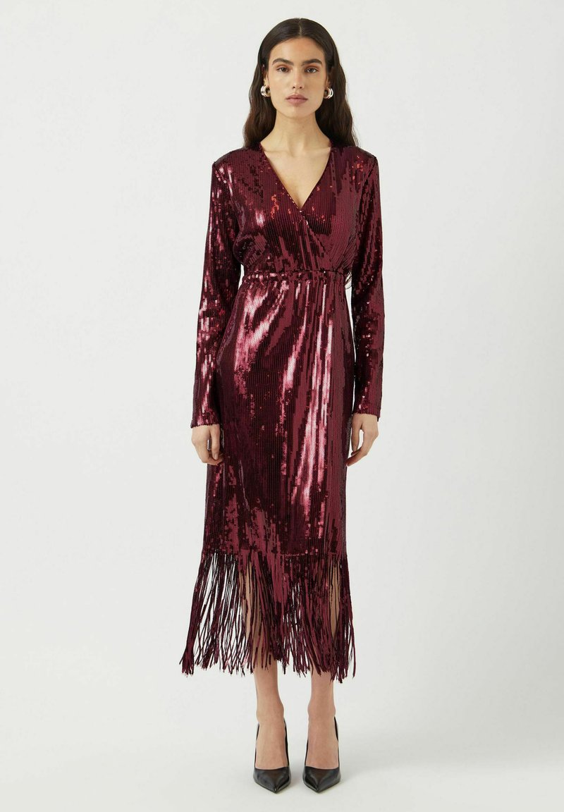 Abito lungo in paillettes bordeaux con scollatura a V, maniche lunghe, vita cin cinata e orlo frastagliato. Abbinato a scarpe nere con tacco alto.