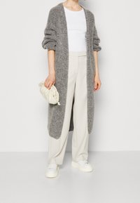 Personne portant un long cardigan gris, un haut blanc, un pantalon beige à jambes larges, des baskets blanches, et tenant une petite pochette beige.