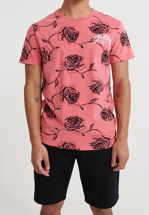 Muž nosící růžové tričko s černými růžovými vzory a bílým logem Superdry, doplněné o černé šortky po kolena.