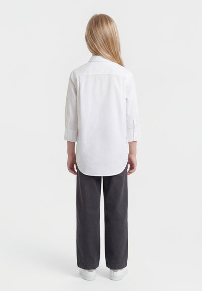 Παιδί με μακριά ξανθά μαλλιά, φοράει λευκό πουκάμισο με κουμπιά, σκούρο παντελόνι με ευρύ πόδι και λευκά αθλητικά παπούτσια, στέκεται με την πλάτη στραμμένη προς τα εμπρός.