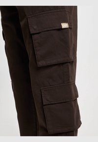 Gros plan sur la jambe d’un pantalon cargo brun foncé avec deux grandes poches à rabat, dont l’une présente une petite étiquette beige portant la mention « DEF ».