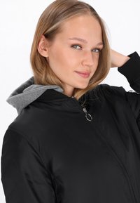 Chaqueta bomber negra con una capucha gris en contraste, puños acanalados y cierre frontal de cremallera. Tela suave y brillante con un diseño clásico.