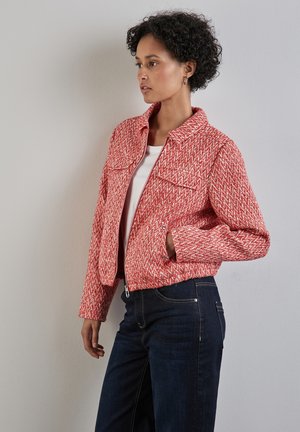 Red and white textured jacket with a zip front, two chest pockets, and a collared neckline, paired with dark denim jeans.