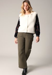Gilet en polaire texturé ivoire avec col rond et fermeture à l'avant, associé à des manches noires brodées de fleurs et un pantalon vert olive.