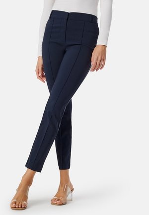 Marineblauwe slim-fit broek met een gladde textuur, voorzien van voornaad, een klassieke tailleband en een getailleerd ontwerp, gecombineerd met doorzichtige haksandalen.