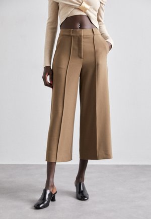Brown, wide-leg trousers with a high waist, featured front pleats, and side pockets, paired with black, open-toe mules.