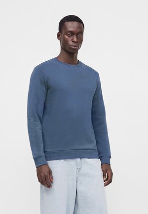 Mand iført en ensfarvet blå langærmet sweatshirt og lyseblå jeans, stående mod en hvid baggrund.