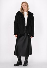 Schwarze Kunstpelzjacke mit großem Kragen, Reißverschluss und Seitentaschen. Kombiniert mit einem langen, schwarzen Satinrock und schwarzen Stiefeletten.