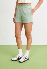 Světle zelené šortky Nike s šedým páskem, které mají kapsy a jemnou texturu. Spárováno s černobíle vzorovanými botami Nike a bílými ponožkami.