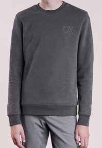 Sudadera gris oscuro de tela suave, con cuello redondo, puños y dobladillo acanalados, y el logo "CK" estampado en el pecho.