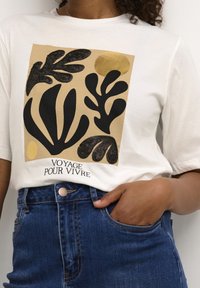 Camiseta blanca con un diseño gráfico que presenta formas botánicas negras y un círculo dorado. El texto dice "VOYAGE POUR VIVRE". Jeans de mezclilla azul de cintura alta.