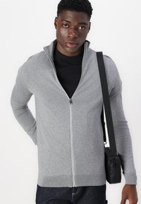 Sweater cinza com zíper, de mangas longas e punhos canelados, usado sobre uma camisa preta. O modelo está segurando uma bolsa preta tiracolo.