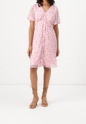 Vrouw in een roze bloemen jurk tot de knie met knopen aan de voorkant en beige vetersandalen met hakken, staand tegen een witte achtergrond.