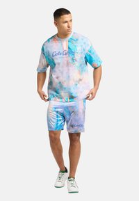 Camiseta gráfica de manga corta azul con patrones abstractos y logo, combinada con pantalones cortos a juego. Zapatillas blancas con acentos verdes.