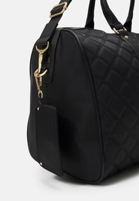 Černá prošívaná kabelka se zlatými detaily, zaobleným tvarem, dvojitými ušima a odnímatelnou kapsičkou na štítek na boku. Hladký povrch.