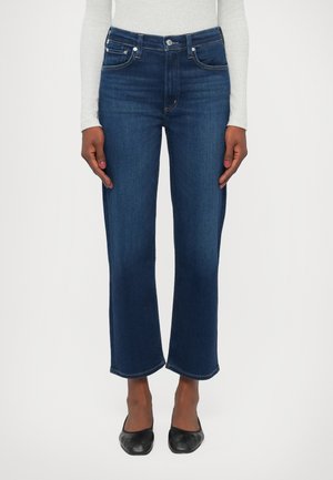 Femme portant un jean droit bleu foncé court, des ballerines noires et un haut à manches longues gris clair, debout devant un fond uni.