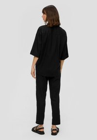 Czarna luźna koszula z krótkim rękawem i przednią listwą, zestawiona z czarnymi spodniami. Noszona z sandałami na odkrytych palcach. Prosty, minimalistyczny styl.