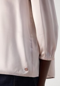 Light pink satin blouse with a gathered sleeve and a silver logo accent near the hem. Smooth texture and loose fit characterize the design.