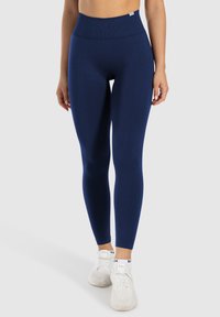 Niebieskie legginsy z wysokim stanem, wykonane z elastycznego materiału, z bezszwowym designem i gładką strukturą, w połączeniu z białymi butami sportowymi.