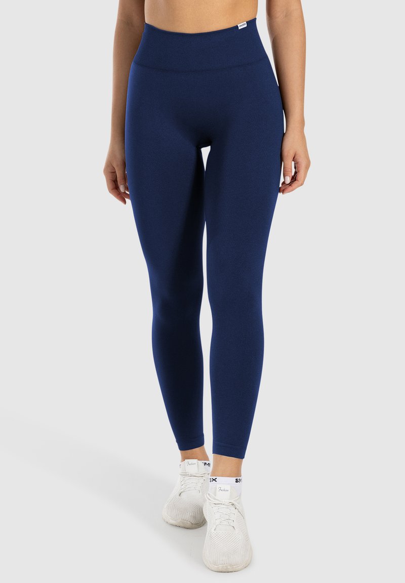 Niebieskie legginsy z wysokim stanem, wykonane z elastycznego materiału, z bezszwowym designem i gładką strukturą, w połączeniu z białymi butami sportowymi.