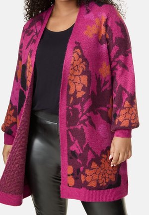 Vrouw draagt een levendige roze cardigan met oranje en donkere bloemenprints over een zwart topje en zwarte broek met leren uitstraling.