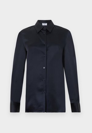 Femme noire debout, les mains dans les poches, portant une chemise en satin bleu marine et un pantalon plissé crème à jambes larges sur fond blanc.