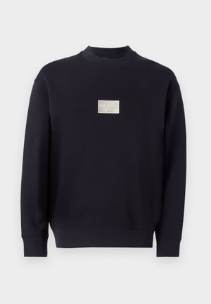 Mørkeblå sweatshirt laget av myk bomull, med rund hals, lange ermer, og en liten sølvfarget logo-patch foran.