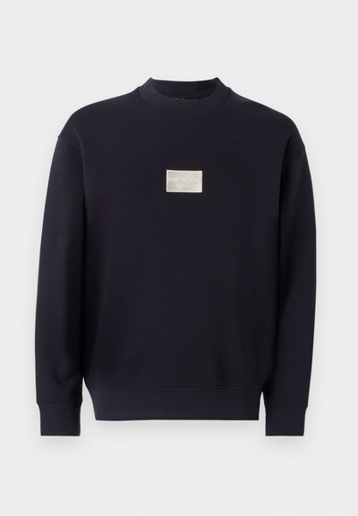 Sweatshirt bleu marine en coton doux, avec un col rond, des manches longues et un petit patch logo argenté à l'avant.