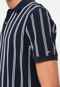 Námořnická polokošile s bílými vertikálními pruhy, texturovaná pletenina, límec na dvě knoflíky a malý logovaný detail na rukávu.