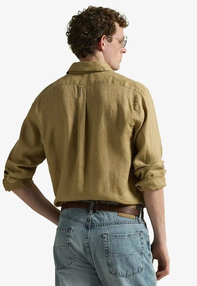 Homme aux cheveux bouclés, chemise beige manches retroussées et jean bleu clair avec ceinture marron, vu de dos sur fond blanc.