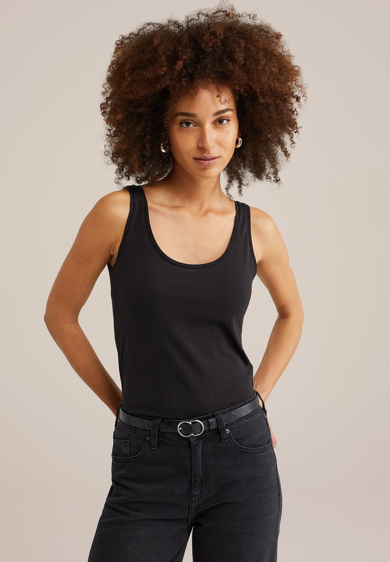 Nainen, jolla on kiharat hiukset, pukeutuneena mustaan hihattomaan paita, mustiin farkkuihin ja mustaan vyöhön, seisoo kädet selän takana.