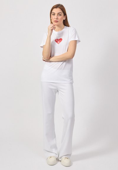 T-shirt bianco in cotone con maniche corte, con una grafica di un cuore rosso, abbinato a pantaloni bianchi a campana e scarpe slip-on di colore chiaro.
