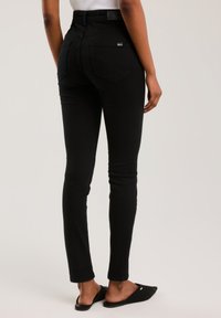 Naine, kes kannab kitsaid musti teksapükse ja musta libisevaid kingi, seisab selg kaamerale vastu tavalise taustaga.