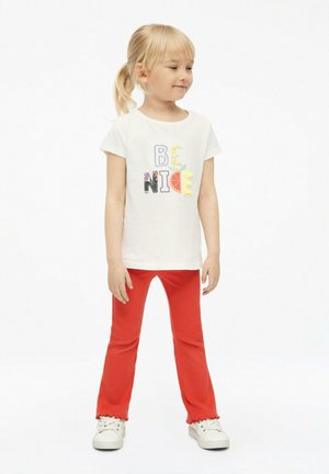 Junges Mädchen mit blondem Pferdeschwanz, das ein weißes "Be Nice"-T-Shirt, rote ausgestellte Hosen und weiße Sneakers trägt, steht vor einem schlichten Hintergrund.