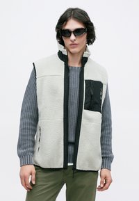 Jovem vestido com óculos de sol pretos, suéter de malha cinza, colete de fleece creme com bolso zwarte com zip, e calças verde-oliva, contra um fundo branco.