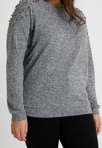 Grijze trui met een gestructureerde stof, voorzien van metalen studs op de schouders, lange mouwen en een ronde halslijn.