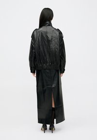 Giacca di pelle nera con vestibilità ampia e orlo elasticizzato, indossata sopra una lunga gonna di pelle nera con uno spacco laterale. Scarpe con tacco alto visibili.