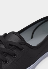 Μαύρο αθλητικό παπούτσι χαμηλού ύψους με υφασμένο δερμάτινο μπροστινό μέρος, μαύρα κορδόνια και λευκή λαστιχένια σόλα, δείχνεται σε κοντινή γωνία.