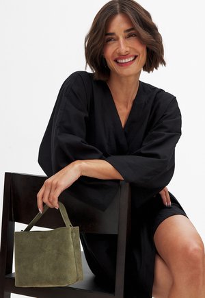 Top in stile kimono nero con maniche larghe. Il modello tiene in mano una piccola borsa verde in suede con una forma strutturata e manico superiore.