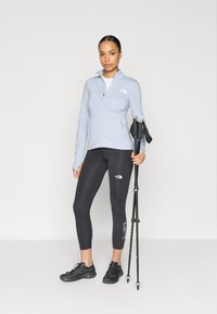 Ανοιχτό μπλε τζάκετ με φερμουάρ, μαύρο κολάν με λογότυπο και μαύρα ορειβατικά παπούτσια. Το μοντέλο κρατά μπαστούνια πεζοπορίας, μινιμαλιστικό φόντο.